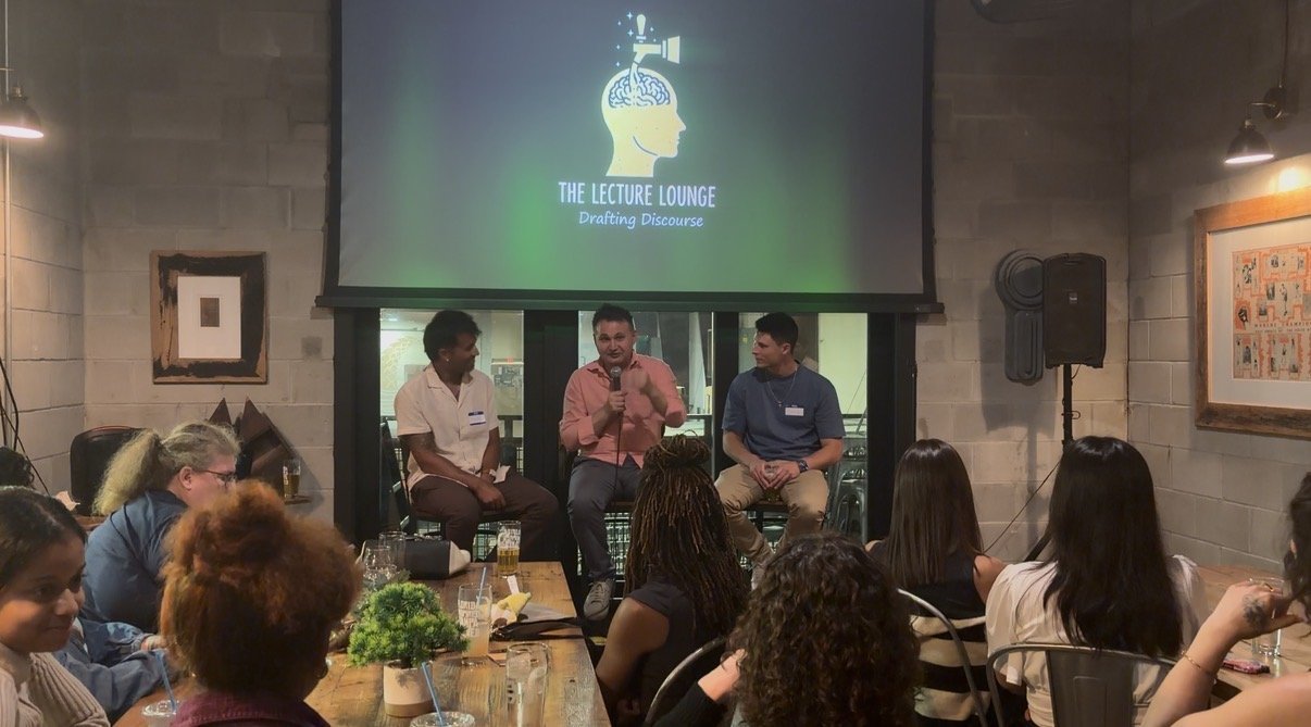 Dr. Razvan Teodorescu on stage at The Lecture Lounge with two co-panelists, the audience seated at a long brewery table in front of a branded projection screen